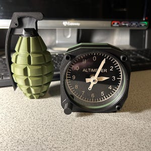 May include: An olive green grenade-shaped object and a black and green altimeter clock. The clock face displays the word "ALTIMETER" and numbers from 0 to 11. The grenade has a black handle and a textured surface.