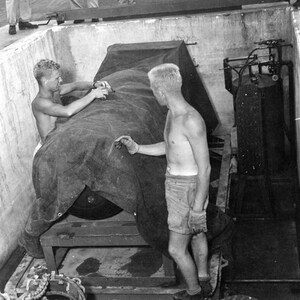 May include: Two men in a black and white photograph are loading a large, covered object onto a flatbed lorry. The object is covered with a tarpaulin and secured with chains. The men are wearing shorts and are shirtless. The photograph is labelled "Top Secret" and "121".