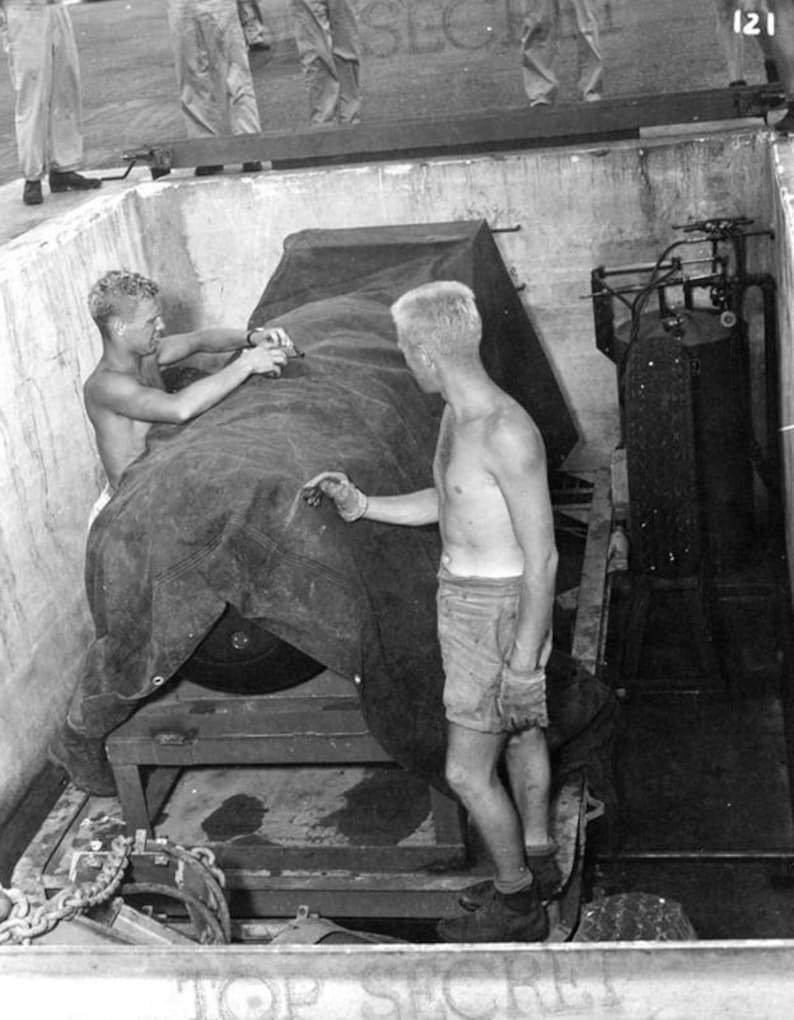 May include: Two men in a black and white photograph are loading a large, covered object onto a flatbed lorry. The object is covered with a tarpaulin and secured with chains. The men are wearing shorts and are shirtless. The photograph is labelled "Top Secret" and "121".