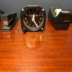 May include: A black and white altimeter clock with a white needle pointing to the number 10. The clock is on a wooden surface with a black stapler, a paperclip, and a black tape dispenser.