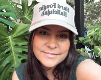 May include: A light-colored trucker hat with the text "Hard Enough Hallelujah" in a dark font. The hat is worn by a person in front of large green plants. The person is wearing a dark green top.