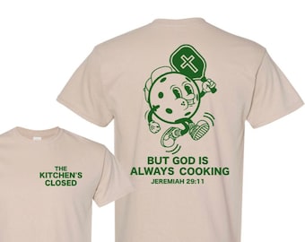 May include: A tan t-shirt with a green graphic of a cartoon character holding a paddle with a cross on it. The text "BUT GOD IS ALWAYS COOKING JEREMIAH 29:11" is printed below the graphic. The front of the shirt has the text "THE KITCHEN'S CLOSED" printed in green.