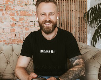 Op de afbeelding: Een man draagt een zwart T-shirt met de tekst "JEREMIAH 29:11" erop gedrukt. Hij zit op een bank en houdt een smartphone vast.