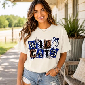 May include: A cream-coloured short-sleeve t-shirt featuring the text WILDCATS in a block-letter style. The letters are filled with various patterns including blue and white checkers, leopard print, and blue polka dots, accented by stars and bows.