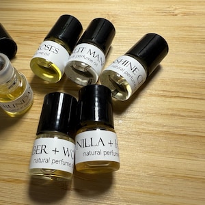 May include: Six small glass bottles with black caps and labels. The labels have white text on them, including the names of the scents: "Roses", "Lit Man", "Shine", "Vanilla + Honey", "Amber + Wood", and "Abundant". The bottles are filled with a clear, light yellow liquid.