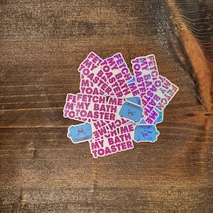 Op de afbeelding: Een verzameling stickers met een blauwe broodrooster met een lachend gezicht. De stickers hebben de tekst "FETCH ME MY BATH TOASTER" in een herhalend patroon. De stickers liggen op een houten oppervlak.