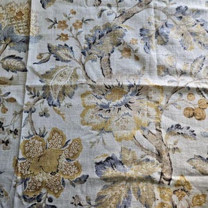 May include: Close-up of a fabric with a floral pattern. The design features large, stylized flowers and foliage in shades of gold, blue, and brown against a cream-colored background. The fabric has a textured appearance.