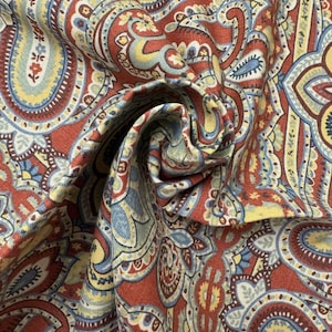 May include: Close-up of a fabric with a detailed paisley pattern. The fabric features a warm color palette of red, yellow, blue, and white. The design includes intricate floral and teardrop shapes, creating a rich, textured appearance. The fabric is folded, showing depth.