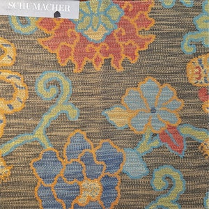 May include: A brown fabric with a floral pattern featuring blue, yellow, orange, and red flowers. The fabric has a textured woven look. The Schumacher logo is in the top left corner.