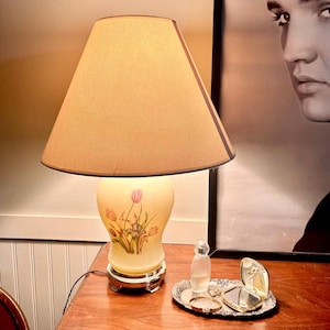 May include: A table lamp with a beige fabric conical shade and a cream-colored glass base featuring a painted floral design of pink tulips and green stems. The lamp sits on a wooden surface next to a small silver tray with a perfume bottle and mirror.
