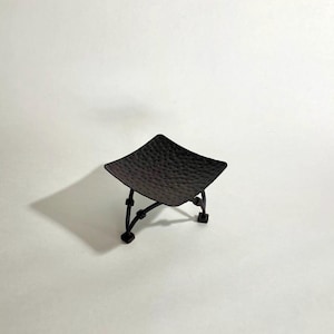 May include: A small, square, black metal dish with a hammered texture, supported by four curved legs. The dish is elevated and appears to be a decorative item, possibly for holding small objects or used as a candle holder.