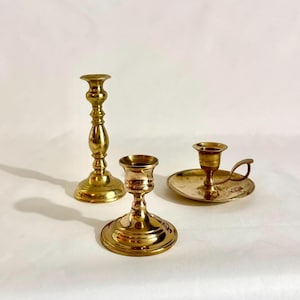 Puede incluir: Tres portavelas de latón pulido con diseños variados. Uno es un candelabro alto y esbelto con base escalonada, otro es un candelabro más corto y ancho, y el tercero es un portavelas bajo con una bandeja circular y un asa lateral.