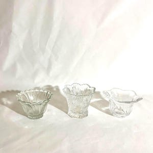 May include: Three clear glass bowls with distinct designs. The left bowl has vertical fluted ribs, the center bowl features a textured diamond and fan pattern with a scalloped rim, and the right bowl has a smooth, wide, wavy-edged rim.