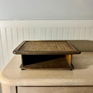 May include: A rectangular wooden display shelf with a woven top. The shelf has a dark brown frame and a light brown woven surface. The shelf is sitting on a light-colored wooden table.