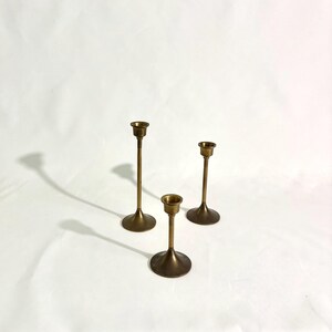 May include: Three brass candlesticks of varying heights stand against a white background. Each candlestick has a circular base, a slender stem, and a small cup-shaped top to hold a candle. The candlesticks are in a warm, metallic tone.