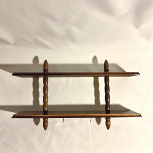 May include: A two-tier wooden wall shelf featuring dark brown horizontal planks connected by four vertical, spindle-shaped supports. The structure includes two small metal hooks attached to the underside of the bottom shelf.