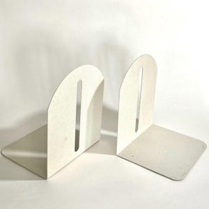 May include: Two off-white metal bookends. Each bookend has a rectangular base and a curved top with a vertical slot. The bookends are designed to hold books upright on a shelf or desk. The bookends are made of a sturdy metal material.