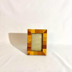 Puede incluir: Un marco de fotos rectangular de madera con un diseño de mosaico. El marco está hecho de varias tonalidades de madera marrón y amarilla. La abertura interior es de color beige claro, adecuada para una foto o una obra de arte.