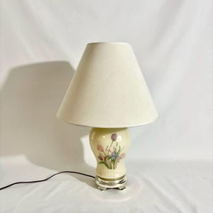 May include: A table lamp with a cream-colored ceramic base featuring a floral pattern of purple and pink tulips and green leaves. The lamp has a tapered, off-white fabric shade and a polished silver-tone metal base with a black power cord.