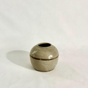 Może przedstawiać: Mały, okrągły, beżowy wazon ceramiczny o gładkim, szkliwionym wykończeniu. Wazon ma szeroki, pękaty korpus, który lekko zwęża się ku wąskiemu, okrągłemu otworowi u góry. Cienka, ciemnobrązowa pozioma opaska otacza środkową część naczynia.