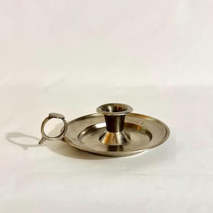 Puede incluir: Un candelabro de metal en tono plateado con una base circular ancha que sirve como bandeja para gotas y un receptáculo central cónico para sostener una vela. Incluye un pequeño asa circular en el lateral para facilitar su transporte.