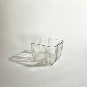 Könnte beinhalten: Eine klare, quadratische Glasschale mit vertikalen Rippendetails. Die Schale hat eine leicht gebogene Form und steht auf einer weißen Oberfläche. Das Glas ist transparent.