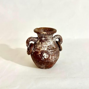 May include: A small, round, brown ceramic vessel with a wide, flared rim and two side handles. Each handle features a circular ring attachment. The surface has a weathered, textured appearance with patches of white, chalky-looking residue.