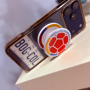 May include: A smartphone case featuring a license plate design with the text BOG-COL and 2024, paired with a circular phone grip displaying a red, yellow, and blue soccer ball graphic with the text FEDERACION COLOMBIANA DE FUTBOL and La Sele.