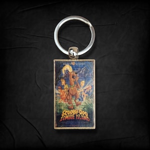 May include: A rectangular keychain featuring the Scooby-Doo Zombie Island movie poster. The image shows Scooby-Doo and the gang in a spooky scene with the text "SCOOBY-DOO ZOMBIE ISLAND". The keychain has a silver keyring.