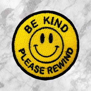 May include: A yellow embroidered patch with a black border. The patch features a smiling face and the text "BE KIND PLEASE REWIND" in black lettering. The background is a marble pattern.