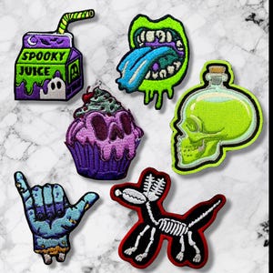 May include: A collection of embroidered patches with a spooky theme. Designs include a juice box with "Spooky Juice" text, a mouth with a blue tongue, a skull cupcake, a skull-shaped potion bottle, a hand with a "hang loose" gesture, and a skeleton dog.