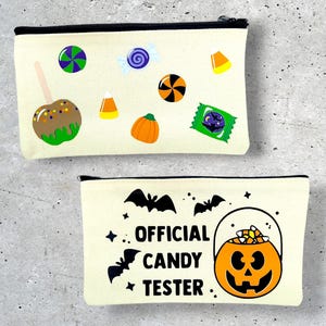 May include: Two canvas zipper pouches with black trim. One features candy and a caramel apple graphic. The other has bats, stars, and a jack-o'-lantern with the text "OFFICIAL CANDY TESTER."