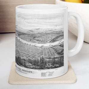May include: A white ceramic mug featuring a detailed black and white historical bird's-eye view illustration of Portland, Oregon. The print includes a river, city grid, and mountains, with the text PORTLAND, OREGON printed at the bottom.