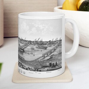 May include: A white ceramic mug featuring a detailed black and white historical illustration of a bustling riverfront city scene with bridges, steamboats, and industrial buildings. The print wraps around the mug, showcasing a vintage panoramic view.