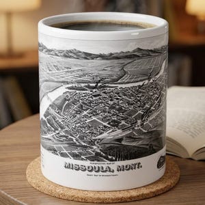 May include: A white ceramic mug filled with dark liquid, featuring a detailed black and white historical perspective map of Missoula, Montana. The text on the mug reads: PERSPECTIVE MAP OF MISSOULA, MONT. COUNTY SEAT OF MISSOULA COUNTY.