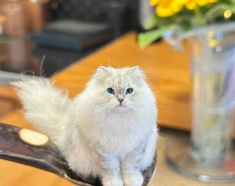 May include: A fluffy, white cat with light blue eyes sits on a decorative spoon. The cat has long fur and a bushy tail. The spoon is dark brown with a gold-colored accent. A vase of sunflowers is in the background.