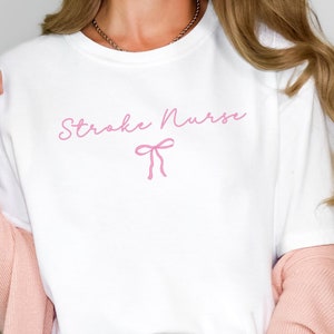 May include: White t-shirt with the text "Stroke Nurse" in pink script with a pink bow underneath.