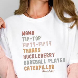May include: White t-shirt with a graphic design featuring the words "Mama", "Tip-Top", "Fifty-Fifty", "Thanks", "Huckleberry", "Baseball Player", and "Caterpillar" in a rainbow colour scheme. The text "#nihhss" is printed below the graphic.