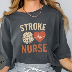 May include: A dark gray t-shirt with the text "STROKE NURSE" in orange and white. A stylized brain and a red heart with a heartbeat graphic are above the text.