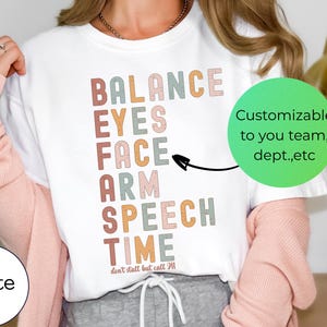 Op de afbeelding: Wit T-shirt met de woorden "BALANCE, EYES, FACE, ARM, SPEECH, TIME" in verschillende kleuren. De tekst "don't stall but call M" staat onder het woord "TIME". Een groene cirkel met de tekst "Customizable to you team, dept.,etc" staat rechts.