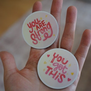 Peut inclure: Deux autocollants ronds roses et blancs avec des messages de motivation. L'autocollant du haut dit "You Are Strong" en lettres roses avec des étoiles jaunes. L'autocollant du bas dit "You Got This" en lettres roses avec des étoiles jaunes et des cœurs roses.