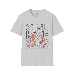 Peut inclure: Un t-shirt gris clair avec un imprimé graphique de deux personnes en combinaisons orange. Le texte "OTSUKARE SAMA DESHITA" est imprimé en lettres noires sur l'image.