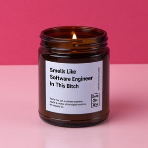 May include: A lit brown glass candle jar with a white label. The label reads "Smells Like Software Engineer In This Bitch." Additional text on the label says "You're not just a software engineer; you're a creator of the digital solutions we depend on."