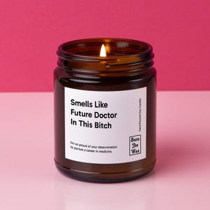 May include: A brown glass candle jar with a white label that reads "Smells Like Future Doctor In This Bitch". The label also includes the text "I'm so proud of your determination to pursue a career in medicine." and the logo "Burn The Wax".