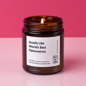 May include: A brown glass candle jar with a white label that reads "Smells Like World's Best Optometrist". The label also includes the text "You help us see the world more clearly, and that's a wonderful gift." and a logo that says "Burn The Wax". The candle is lit and the flame is visible.