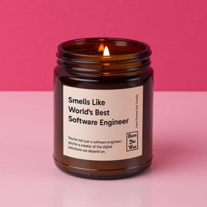 May include: A lit brown glass jar candle with the text "Smells Like World's Best Software Engineer." The candle has a white label with additional text. The background is a solid pink color.