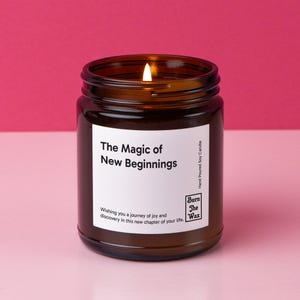May include: A lit soy candle in a brown glass jar. The label reads "The Magic of New Beginnings" with additional text. The candle is hand-poured and made by "Burn the Wax". The background is a solid pink.