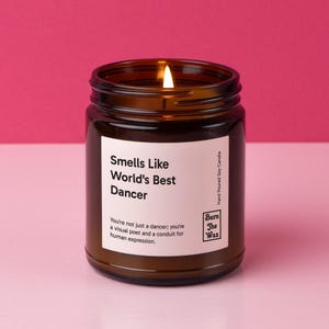 May include: A brown glass candle jar with a white label that reads "Smells Like World's Best Dancer". The label also includes the text "You're not just a dancer; you're a visual poet and a conduit for human expression." and the logo "Burn The Wax".