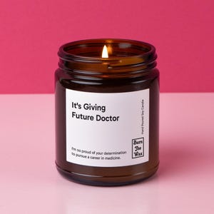 May include: A lit soy candle in a brown glass jar. The white label reads "It's Giving Future Doctor." Below the text is a message of encouragement. The candle is hand-poured and has a burn time of approximately 50 hours.