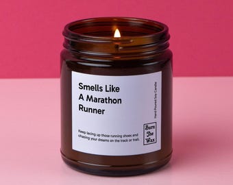 May include: A brown glass candle jar with a white label that reads "Smells Like A Marathon Runner". The label also includes the text "Keep lacing up those running shoes and chasing your dreams on the track or trail." and the logo "Burn The Wax" with the text "Hand Poured Soy Candle".